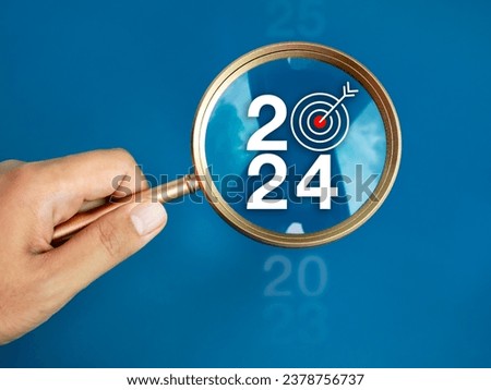 Trends searching, business goals and planning and Happy New Year 2024 concepts. The big white 2024 year number with target icon inside the gold magnifying glass holding by hand on blue background.