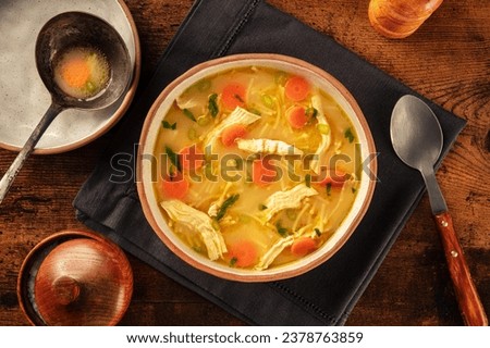 Chicken noodle soup with vegetables, a bowl of healthy broth, overhead flat lay shot on a rustic wooden table, winter home cooking