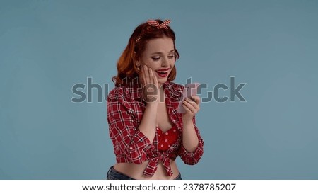 In the picture on a blue background, a young woman with bright makeup. Looks at the phone. Maybe talking to someone or something is watching. Depicts joy, delight, tenderness. HDR BT2020 HLG Material.