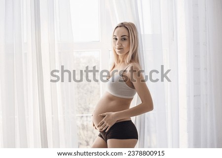 A young blonde pregnant woman stands by the window and strokes her belly in the rays of the morning sun. tender photo session of the expectant mother. pregnant woman in a short top and shorts
