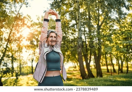 Active aging and outdoor workout. Enduring mature woman with grey hair doing regular workout on fresh air. Positive lady wearing sport outfit performing arms exercises and strengthening back. Royalty-Free Stock Photo #2378839489