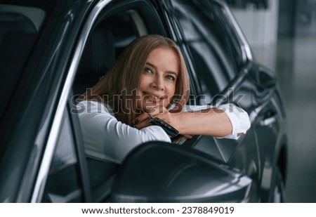 Looking from the opened window. Woman is sitting in a car and driving it.