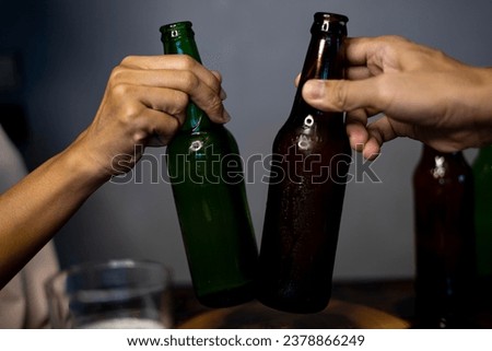 Two Friends Toasting With Bottle Of Light Beer. Hands lifting two beer bottle and happy enjoying harvest time together to clinking glasses at indoor party
