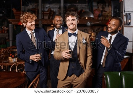 bachelor party, happy interracial best men looking at groom in suit standing with whiskey in bar Royalty-Free Stock Photo #2378883837