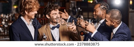 happy friends in formal wear congratulating groom in bar, interracial men holding whiskey banner Royalty-Free Stock Photo #2378883875