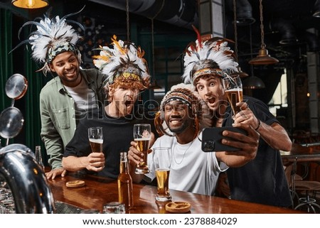 happy multicultural men in headwear with feathers taking selfie on smartphone during bachelor party Royalty-Free Stock Photo #2378884029