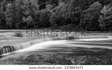 Panoramic view of a weir at Nachrodt-Wiblingwerde in Sauerland Germany. Artifical concrete walls damming up river lenne after heavy rain. Black and white waterfall scenery with longtime exposure.  Royalty-Free Stock Photo #2378896471