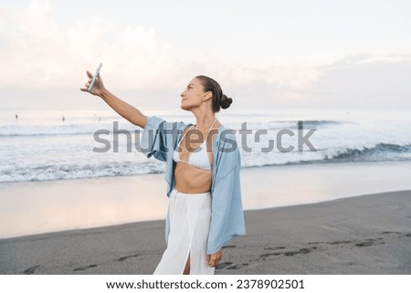 Side view of young female in casual clothes standing on sandy beach and taking selfie on mobile phone while looking at screen and enjoying summer vacation near sea during daytime