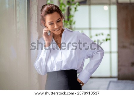 Portrait of a brown haired middle-aged woman wearing a white shirt and black pants and standing by the window. Attractive female cheerful smiling. 