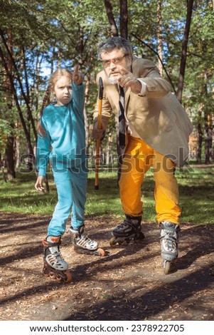 A girl roller skates in the park with her old grandfather. A funny grandfather and a little girl are playing and joking. A grandfather on roller skates plays pranks with his granddaughter.