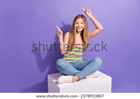 Full size photo of pretty teenager blonde girl sit cube point empty space wear trendy striped outfit isolated on purple color background