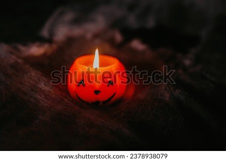 Halloween at home, room decoration ideas. A wax candle in the shape of a smiling pumpkin in a spider web. Symbol of All Saints' Day, October holidays. Natural candle flame. Dark background.