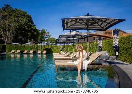 Young woman in white sundress lounges on pool chair at tropical resort, soaking up sun and savoring vacation. The turquoise waters shimmer, lush greenery and picturesque skies surround her