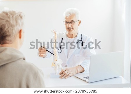 Middle aged male doctor pointing on human spine model with a pen and explaining patient his problem in medical clinic. Spinal healthcare and back pain treatment concept. Selective focus, copy space Royalty-Free Stock Photo #2378955985