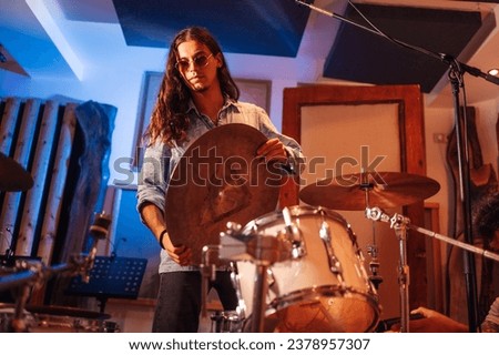 Picture of a young talented drummer with long hair setting up a ride cymbal onto his ivory drum set. Stylish, minimalist music studio. Preparation for music recording session in an isolation room.