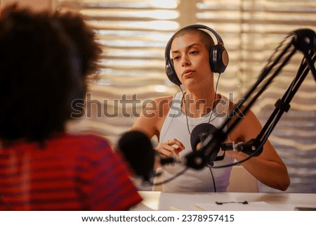 A gen z girl is sitting in a home broadcasting studio with headphones on and speaking on the mic while an interracial interviewer is sitting across the table in a blurry foreground. Girls podcasting.