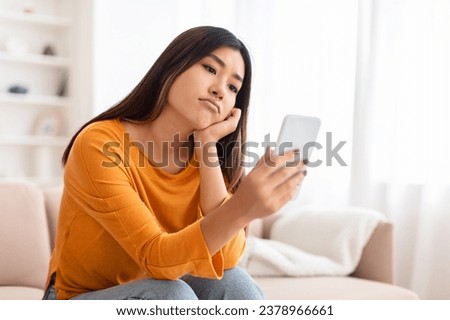 Smartphone Addiction And Depression. Frustrated bored asian young lady using mobile phone, checking social media, reading bad news sitting on the couch. Upset woman waiting for his call, copy space Royalty-Free Stock Photo #2378966661