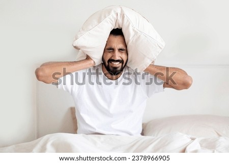 Stressed Young Indian Man Sitting In Bed And Covering Ears With Pillow, Annoyed Eastern Man Feeling Irritated By Loud Noise While Resting In Bedroom, Having Sleeping Problems, Suffering Insomnia Royalty-Free Stock Photo #2378966905