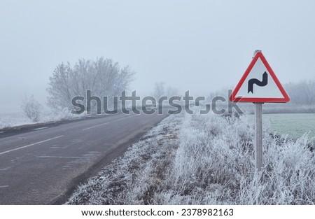 Winter morning on a winding road. The trees and ground are covered in a thick layer of frost. The road turning around a bend. A signpost is standing in front of the bend, warning of a sharp turn. Royalty-Free Stock Photo #2378982163