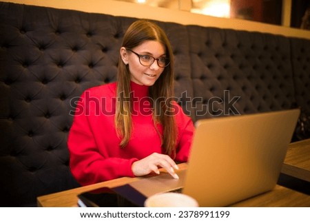 Young female in glasses and warm red sweater professional resume writer checking e-mail via laptop computer while sitting in coffee shop during free time in autumn day 
