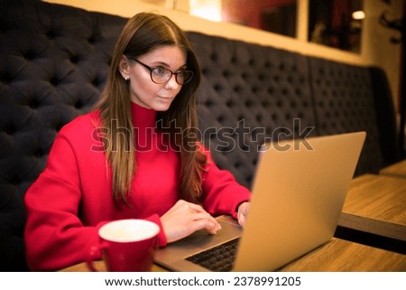 Woman in glasses and warm red sweater skilled social media manager reading notifications on laptop computer while sitting in coffee shop during work break 