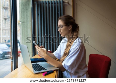 Woman in glasses skilled blogger received notifications on mobile phone during distance work on laptop computer while sitting in coffee shop. Female checks the status of the account on smartphone 
