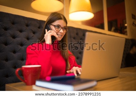Business woman skilled marketing coordinator having smartphone conversation during online work on laptop computer while sitting in restaurant interior 