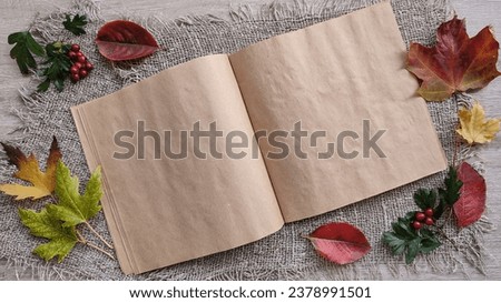 A drawing notebook made of kraft brown paper is unfolded. Clean pages. Framed with colored fallen leaves and wild hawthorn berries. Set against a rough linen background. Close-up. Autumn concept