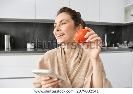 Young woman thinks what to cook, sits in the kitchen with smartphone and tomato in hands, looks aside and smiles, searches recipes on mobile app, orders groceries.