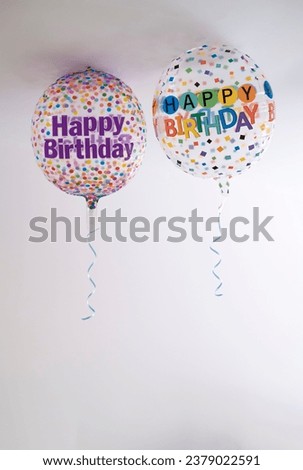 two helium balloons decorated with colored dots and a happy birthday message, with a hanging ribbon, on the roof of a white room, copy space, vertical