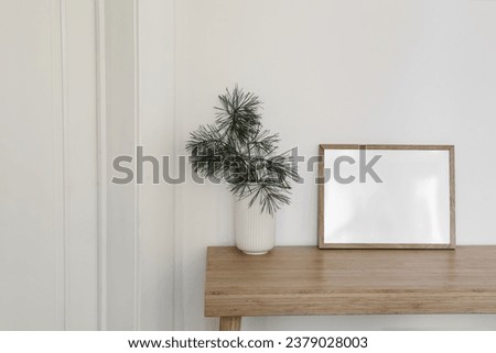 Christmas Scandinavian interior. Minimal winter artistic composition. Blank horizontal wooden picture frame mockup. Pine tree branches in vase on table, desk. White wall background. Empty copy space. 