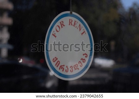 Street photo. City. Round white sign with blue line and red letters on glass door. Shop window. Concept of real estate agency, commercial premises, store showcase. Translation from Georgian: For rent