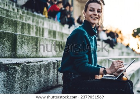 Portrait of cheerful hipster girl with electronic stylus and portable pc resting at city historic center