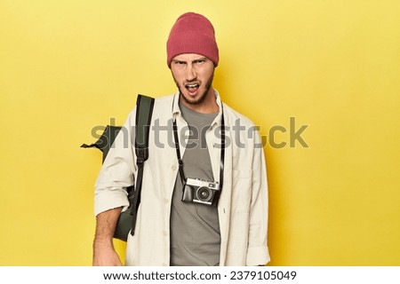 Man with vintage camera and backpack on yellow screaming very angry and aggressive.