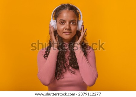 Young beautiful casual Indian woman in wireless headphones looks at camera listening to audiobook or online educational podcast with useful information stands on plain orange background.