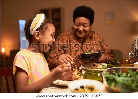 Grandmother putting salad in plate of joyful little girl at family dinner