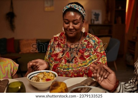 Smiling Black woman in traditional clothes praying before family dinner with Kwanzaa