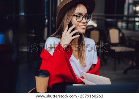 Pleased hipster woman in hat talking on smartphone while working at table on cold cafe terrace