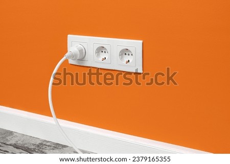 White triple outlet installed on orange wall with inserted white electrical plug, side view. Royalty-Free Stock Photo #2379165355