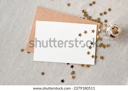 Blank paper card mockup and envelope on neutral beige linen tablecloth with gold star confetti scattered from bottle. Minimalist aesthetic New Year, birthday postcard or invitation template.
