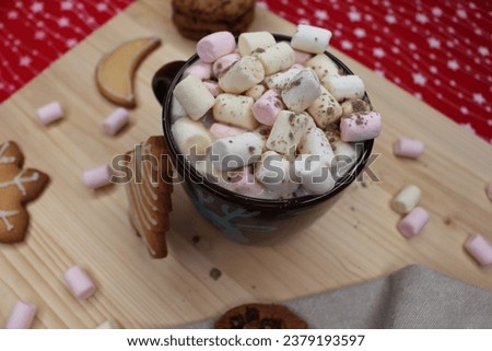 Brown cup or mug of coffee or hot chocolate with marshmallows and cocoa powder. Cookies with chocolate chips and with Christmas shapes. A gray kitchen rag. A light wood and a red and white tablecloth.