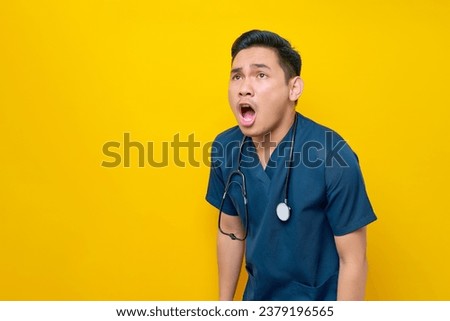 Surprised professional young Asian male doctor or nurse wearing a blue uniform looking aside and open mouth isolated on yellow background. Healthcare medicine concept