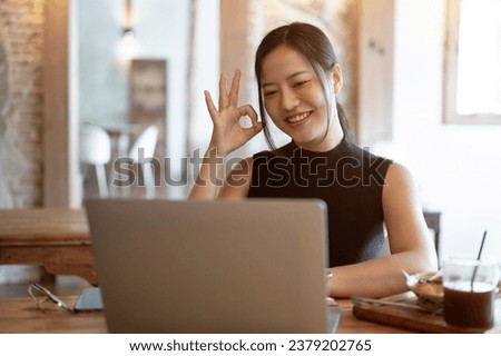 A beautiful Asian businesswoman is joining an online meeting with her team while working remotely at the coffee shop, showing the Okay hand sign. Good, correct, confirm, deal, agreement