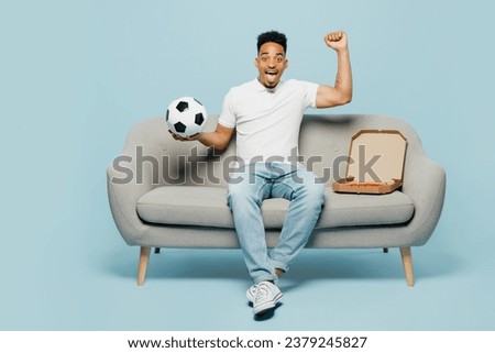 Full body excited happy fun young man fan wear t-shirt cheer up support football sport team hold soccer ball sit on grey sofa do winner gesture watch tv live stream isolated on plain blue background Royalty-Free Stock Photo #2379245827