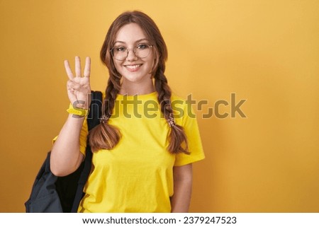 Young caucasian woman wearing student backpack over yellow background showing and pointing up with fingers number three while smiling confident and happy. 