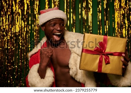overjoyed african american man in santa costume with christmas gift under snow in shiny studio