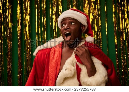 amazed african american man in santa costume with open mouth and christmas bag near golden tinsel