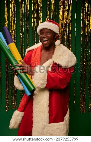 excited african american man in santa costume with colorful wallpapers on green backdrop with tinsel