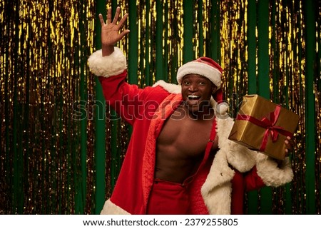 jolly african american man in christmas costume holding present and waving hand on shiny backdrop