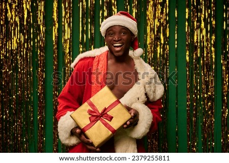 happy african american man in santa costume on shirtless body with shiny gift box near golden tinsel
